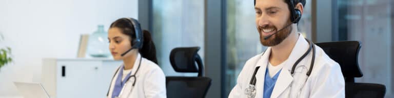 Two medical professionals wearing headsets and white coats providing telehealth support from an office setting.