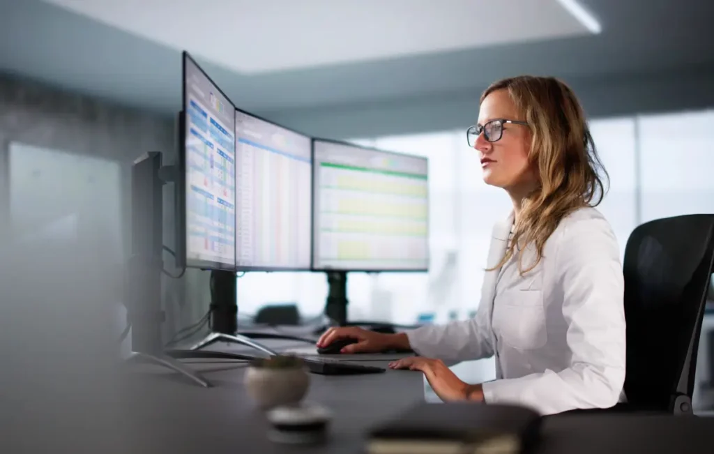 Female healthcare professional in white coat reviewing medical data spreadsheets on dual curved monitors at a clinical workstation.