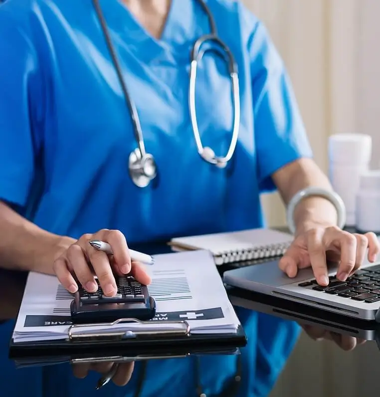 Healthcare professional using a calculator and laptop to review financial documents on a desk.